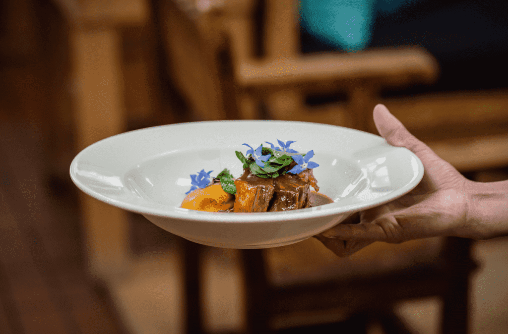 Una mano sostiene un plato blanco de carne cubierto de hierbas y flores de colores.