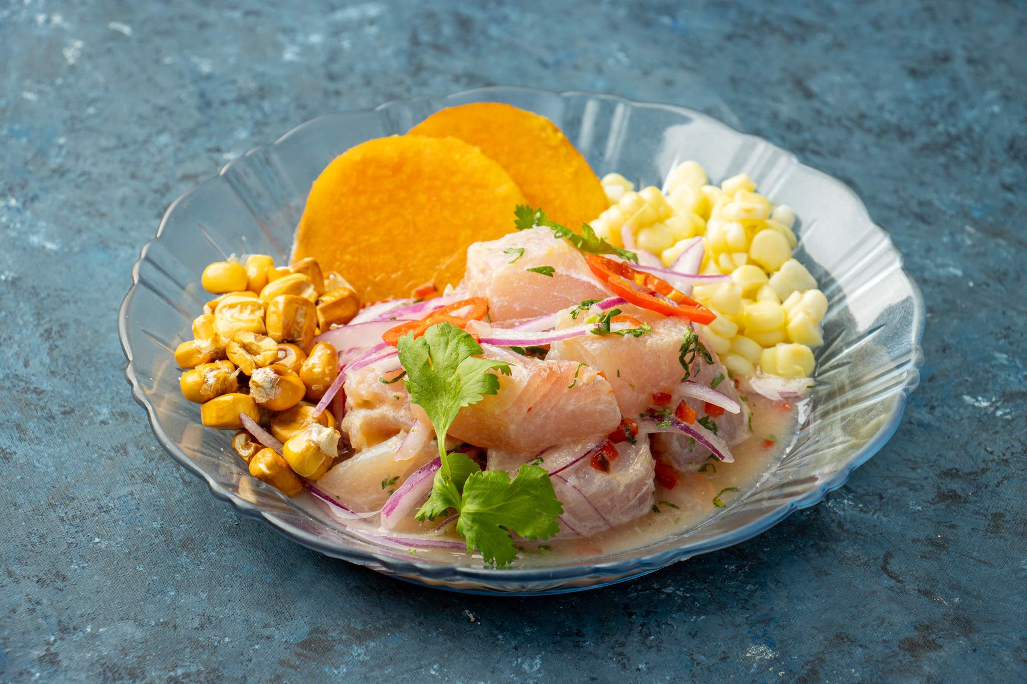 Sobre una mesa azul, un cuenco de ceviche con pesca del día, maíz cancha y fresco y cilantro.