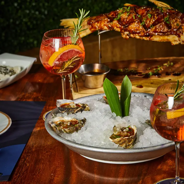 Un plato con ostiones en hielo, copas de vino y al fondo un pescado a las brazas en Fisher’s, uno de los mejores restaurantes de mariscos en CDMX.