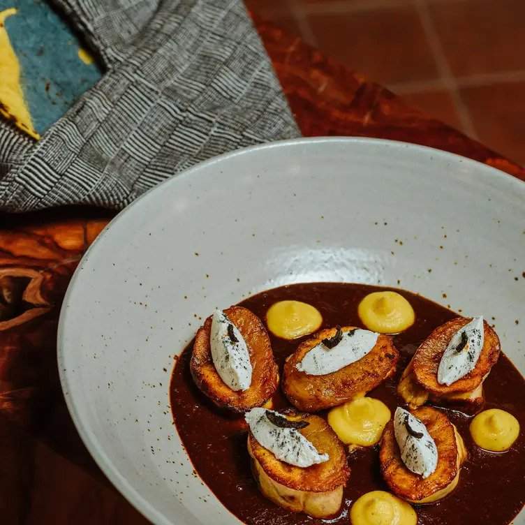 Tazón blanco que contiene un platillo preparado con puré de plátano con una rebanada de plátano macho y queso de cuadro sobre un mole coleto.