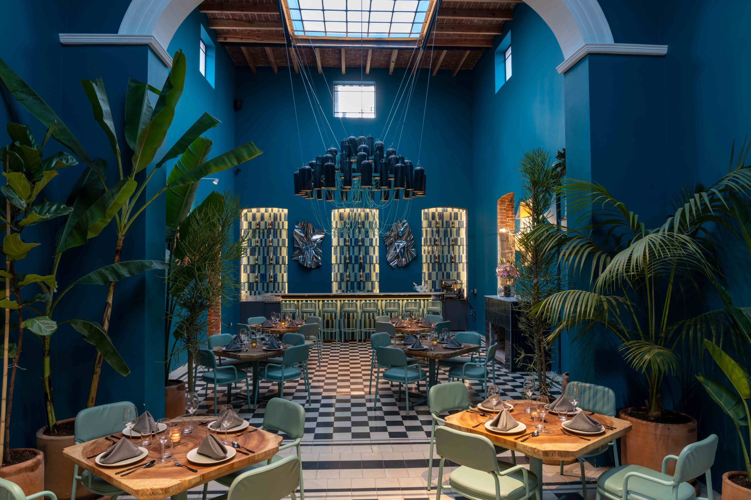 Interior del restaurante Magda, en la Ciudad de México, con muros azules, plantas altas y lámpara central.