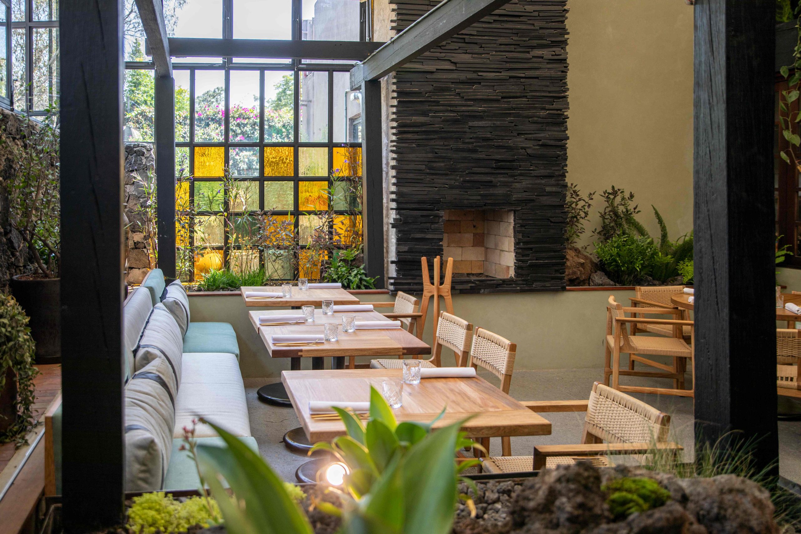 Mesas y sillas de madera claras en el restaurante Sud777 con vitrales de color y piedra volcánica en los muros.