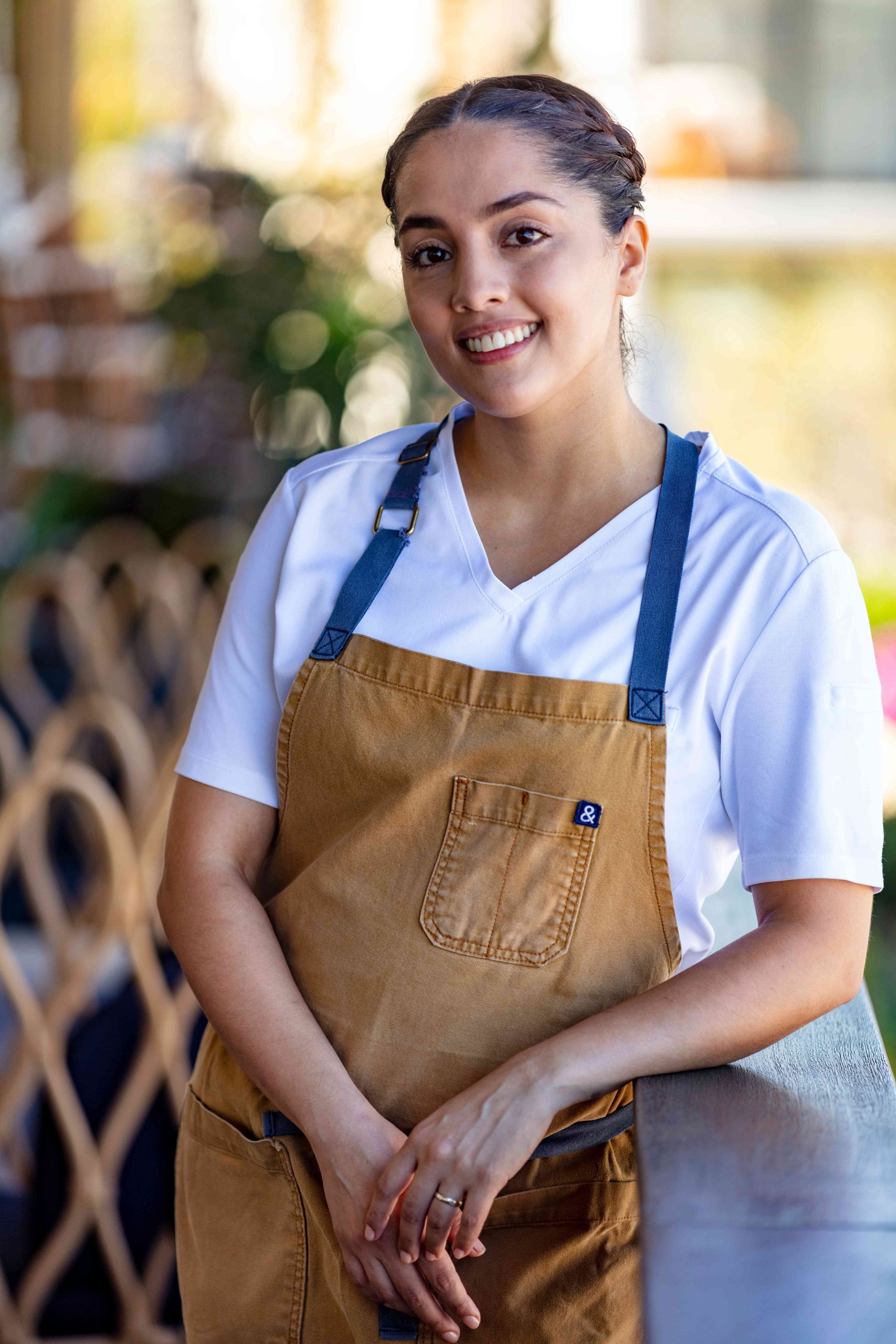 La chef Eliana Godinez sonriendo a la cámara con un delantal color mostaza, posando para la foto para el Dia del chef