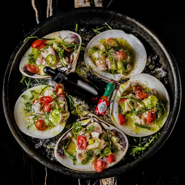 Varias almejas sobre un plato, preparadas con diversas verduras y acompañadas de salsas, en Colmillo.