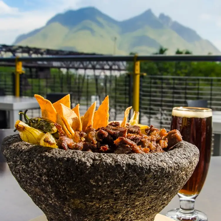 Un molcajete con carne, totopos y una cerveza en una mesa con vista a una montaña en El Lingote, una de las mejores opciones para comer en Monterrey