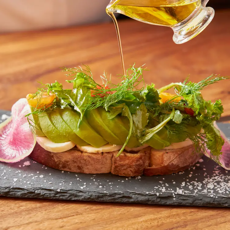 Un platillo de Bencomo, uno de los mejores restaurantes para desayunar en CDMX, donde se ve un pan tostado con rebanadas de aguacate, jitomates cherry y ensalada, que es rociado con un chorro de aceite que cae desde un recipiente.