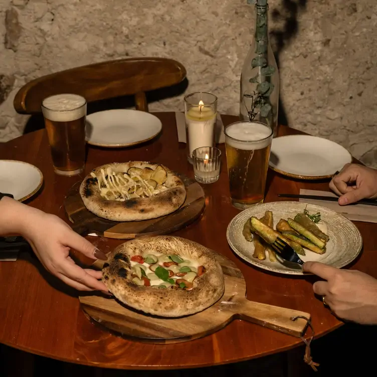 Una mesa con dos tablas de pizza, vegetales, una vela encendida y platos para una pareja comensal.