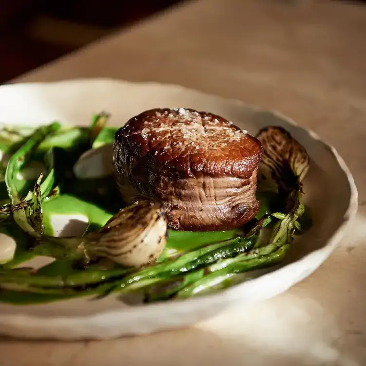 Un plato con un trozo de carne en una cama con diversos vegetales verdes y cebolla asada en Néctar, una de las mejores opciones para comer en Mérida.