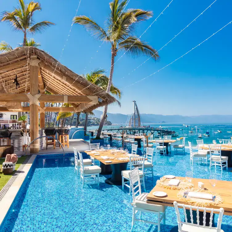 Vista de la alberca y la orilla del mar con mesas cafés con sillas blancas en Mar Y Vino, uno de los mejores restaurantes de Puerto Vallarta.