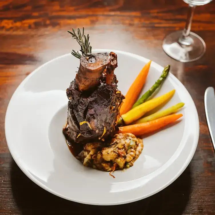 Un platillo de carne acompañado de vegetales sobre un plato blanco y mesa de madera con una copa en La Cappella.